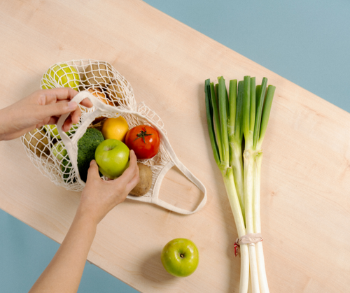 A hand holding a mesh bag with apples, avocados and a bunch of leek on the side